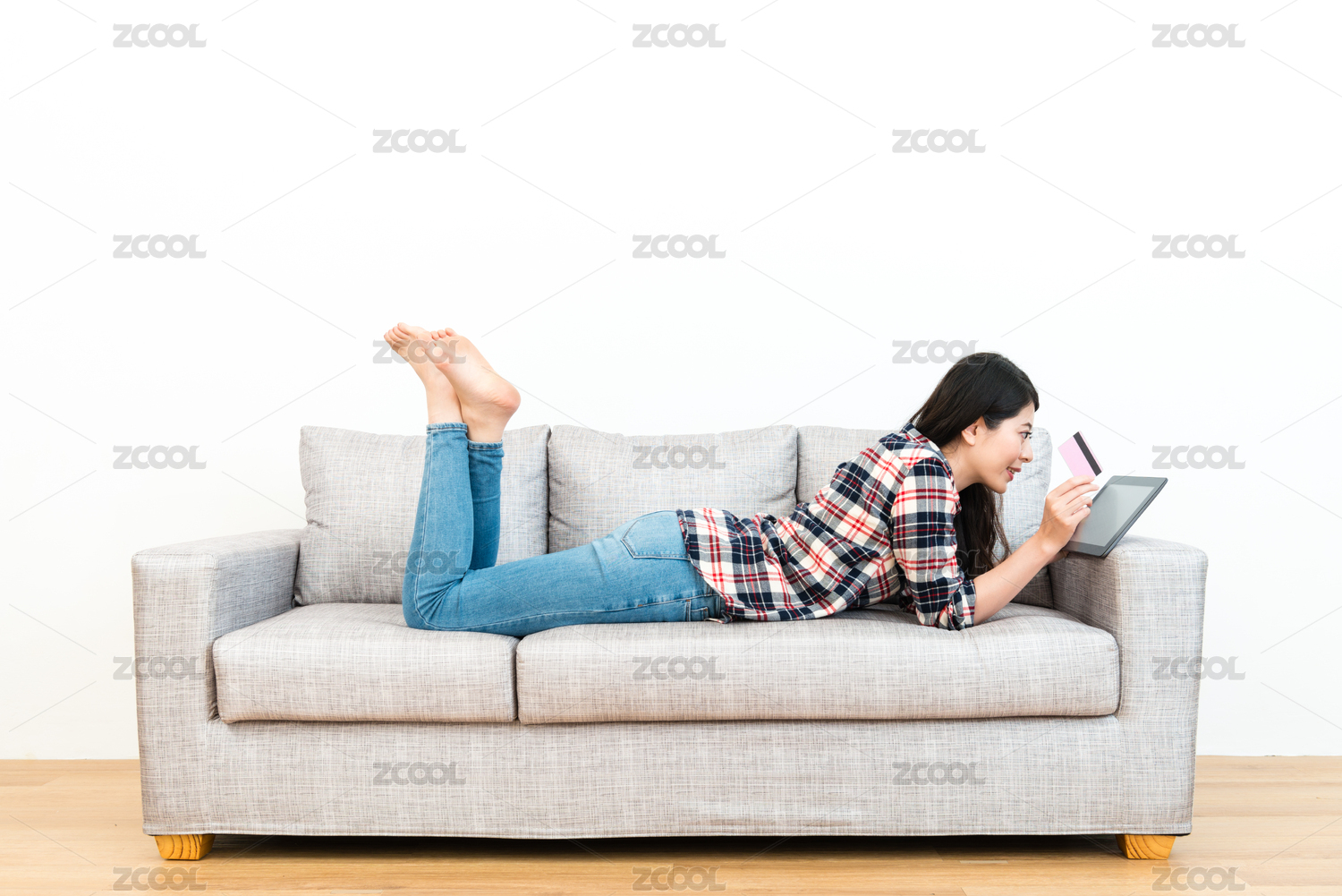 幸福美丽的姑娘在木地板沙发看移动数字平板垫浏览购物网站，并使用个人信用卡的电子商务支付。