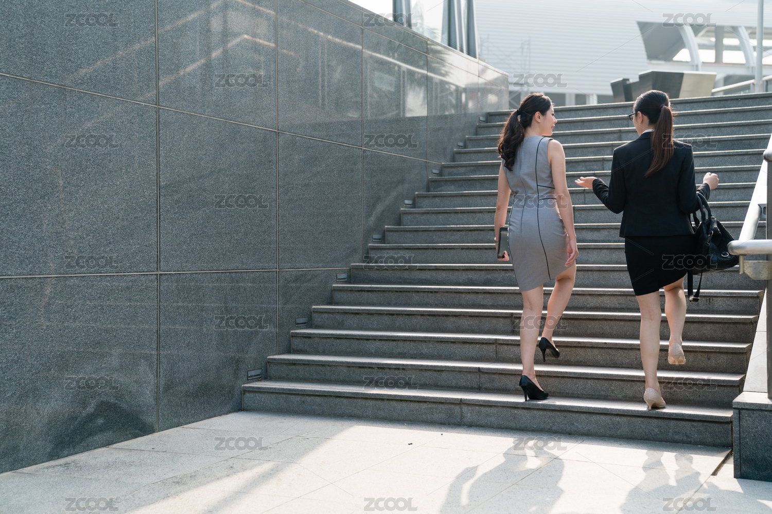 两次成功的亚洲女性走在楼梯上,并与各other.On的后视图聊天。他们要参加一个会议。两人穿着正式西装,高高的山岗。一是平板电脑携带一个手提袋。